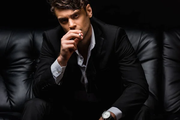 Homme confiant en costume fumer cigarette et regarder la caméra isolé sur noir — Photo de stock