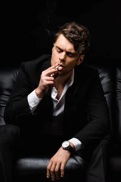 Confident man smoking cigarette and sitting on couch isolated on black — Stock Photo