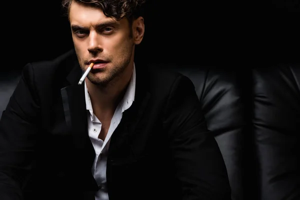 Confident man in formal wear smoking cigarette and looking at camera isolated on black — Stock Photo