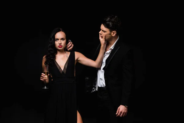 Mujer sexy en vestido sosteniendo copa de champán mientras toca la cara del hombre en traje aislado en negro - foto de stock