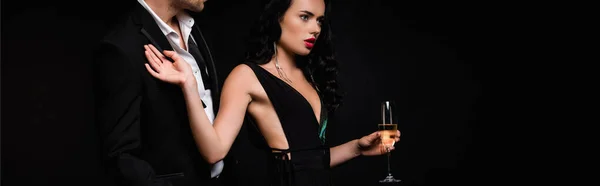 Morena mujer en vestido sosteniendo copa de champán y tirando de hombre en traje aislado en negro, bandera - foto de stock