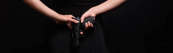 Partial view of dangerous woman holding gun behind back isolated on black, banner — Stock Photo