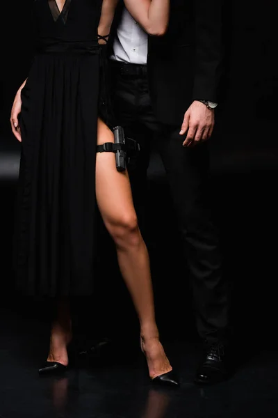 Ausgeschnittene Ansicht einer gefährlichen und bewaffneten Frau im Kleid, die neben einem Mann im Anzug auf schwarz steht — Stockfoto