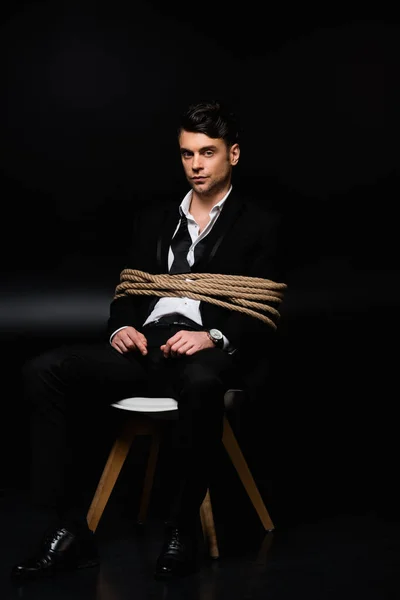 Tied man in suit sitting on chair and looking at camera on black — Stock Photo