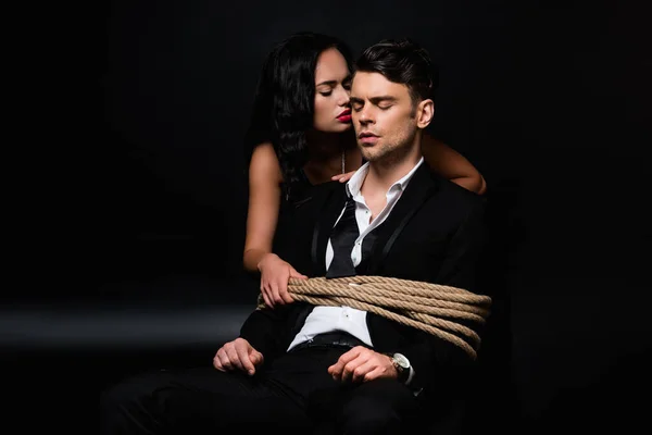 Dominant woman in dress looking at tied submissive man sitting on chair on black — Stock Photo