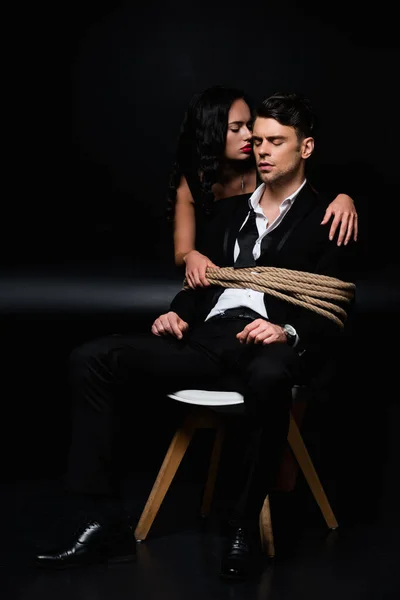 Brunette woman in dress near tied submissive man sitting on chair on black — Stock Photo