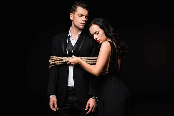 Dominant woman in dress standing with tied submissive man in suit isolated on black — Stock Photo