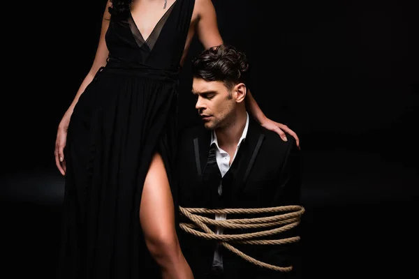 Woman in dress standing with tied submissive man in suit on black — Stock Photo