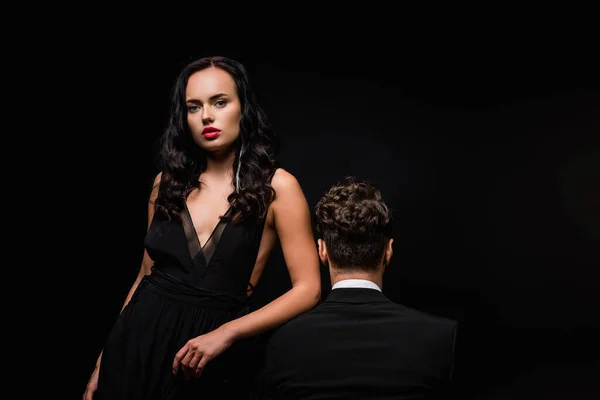 Соблазнительная женщина в платье глядя на камеру рядом с мужчиной в костюме, изолированном на черном — стоковое фото