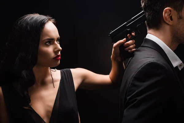 Mujer peligrosa en vestido sosteniendo pistola cerca de hombre en traje aislado en negro - foto de stock