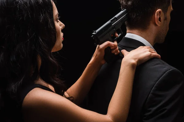 Mujer peligrosa sosteniendo arma detrás del hombre en traje aislado en negro - foto de stock