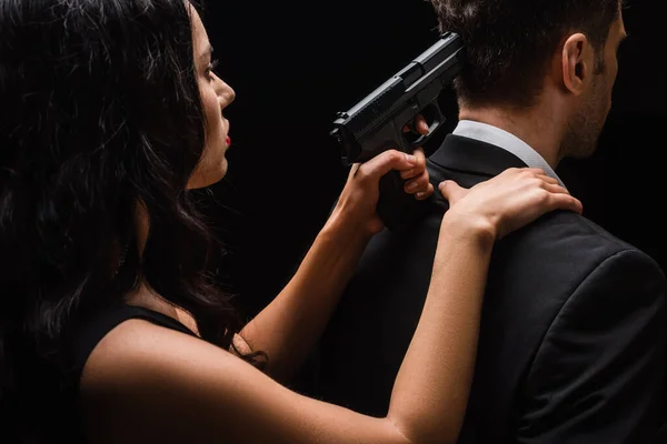 Mujer peligrosa sosteniendo arma detrás del hombre en traje aislado en negro — Stock Photo