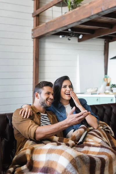 Homme joyeux pointant avec la main près de rire femme couvrant la bouche tout en regardant la télévision sous plaid couverture — Photo de stock