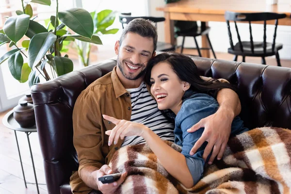 Donna ridente che punta con il dito mentre guarda film vicino sorridente uomo sotto coperta calda — Foto stock