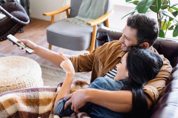 Fröhlicher Mann hält Fernbedienung in der Nähe brünette Frau zeigt mit dem Finger, während Film zu Hause — Stockfoto