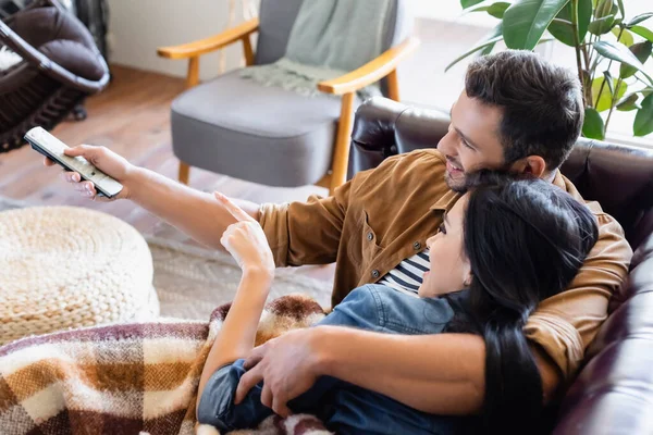 Cheerful man holding remote controller near brunette woman pointing with finger while watching movie at home — Stock Photo