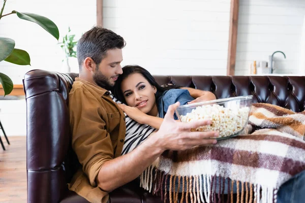 Jovem homem segurando tigela de pipoca enquanto abraçando namorada assistindo tv sob manta xadrez — Fotografia de Stock