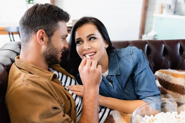 Homem feliz alimentando namorada sorridente com pipocas enquanto descansa no sofá em casa — Fotografia de Stock