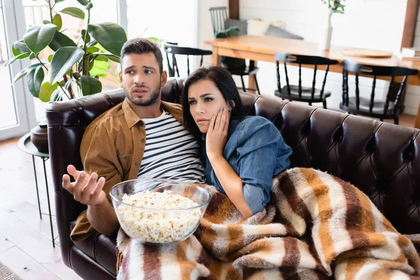 Worried man pointing with hand near tense woman touching face while watching tv near bowl of popcorn — Stock Photo