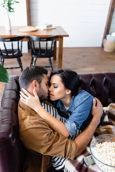 Young couple hugging and kissing on leather sofa near wooden table and chairs on blurred foreground — Stock Photo