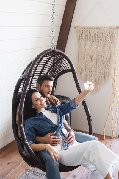 Feliz joven pareja sentado en colgando silla de ratán y tomando selfie en el teléfono móvil - foto de stock