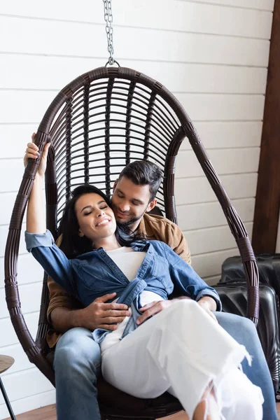 Feliz mujer riendo con los ojos cerrados mientras está sentado con su novio en la silla de ratán colgante - foto de stock