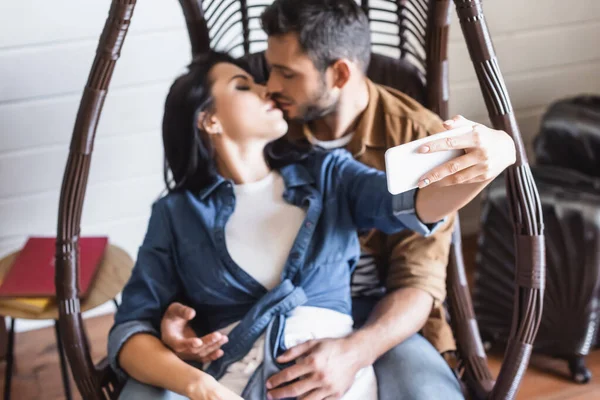 Молодая женщина делает селфи на смартфоне, целуясь с парнем в висящем кресле, размытый фон — стоковое фото
