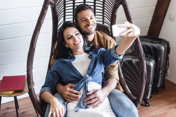 Couple joyeux prenant selfie sur smartphone tout en étant assis dans une chaise en rotin suspendu — Photo de stock