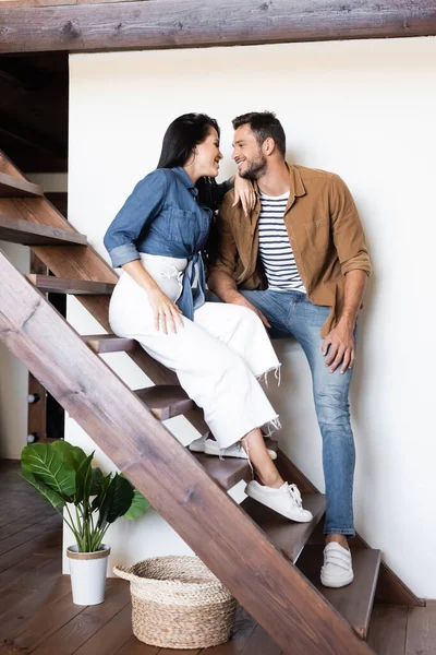 Jovem casal em roupas casuais elegantes olhando uns para os outros em escadas de madeira em casa — Fotografia de Stock