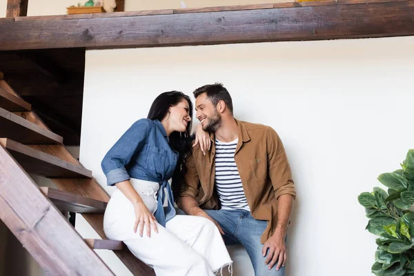 Happy couple in stylish casual clothes looking at each other on wooden stairs at home — Stock Photo