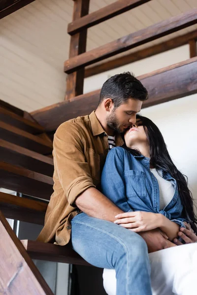 Jeunes amants en vêtements décontractés s'embrassant assis sur des escaliers en bois à la maison — Photo de stock