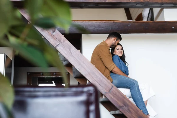Junges Paar umarmt sich, während es zu Hause auf Holztreppen sitzt, verschwommener Vordergrund — Stockfoto