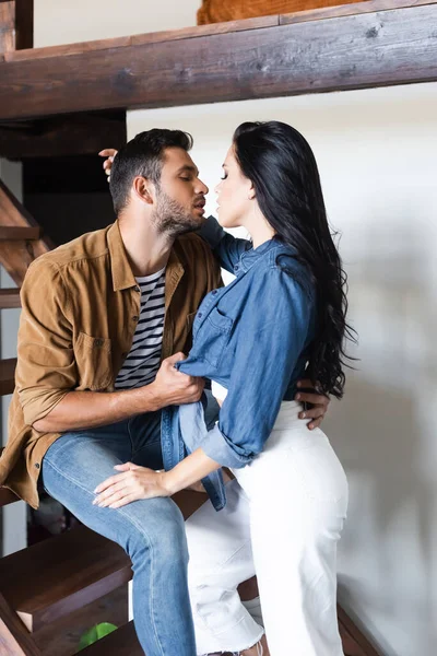 Joven elegante pareja besándose en madera escaleras en casa con los ojos cerrados - foto de stock