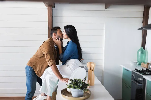 Vista lateral de la mujer en ropa casual con estilo besar a un hombre joven mientras está sentado en el mostrador de cocina - foto de stock