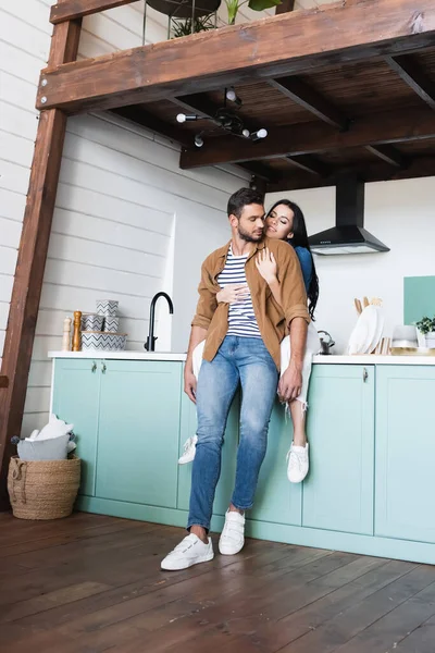Smiling brunette woman embracing young boyfriend while sitting on kitchen counter — Stock Photo