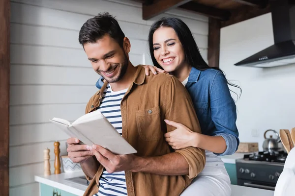 Ridendo donna abbracciando braccio di allegro fidanzato lettura libro in cucina — Foto stock
