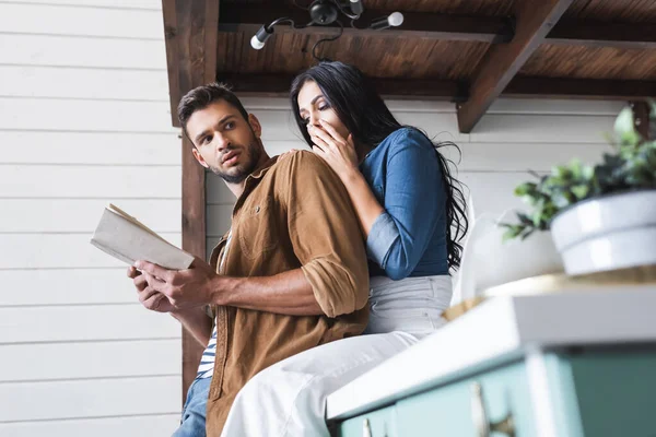 Jeune homme tenant livre près choqué copine couvrant la bouche avec la main — Photo de stock