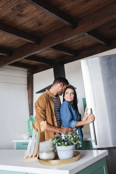 Young man seducing smiling girlfriend opening fridge in kitchen — Stock Photo