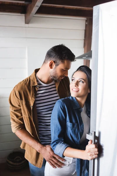Улыбающаяся женщина открывает холодильник и смотрит на мужчину, соблазняющего ее на кухне — стоковое фото