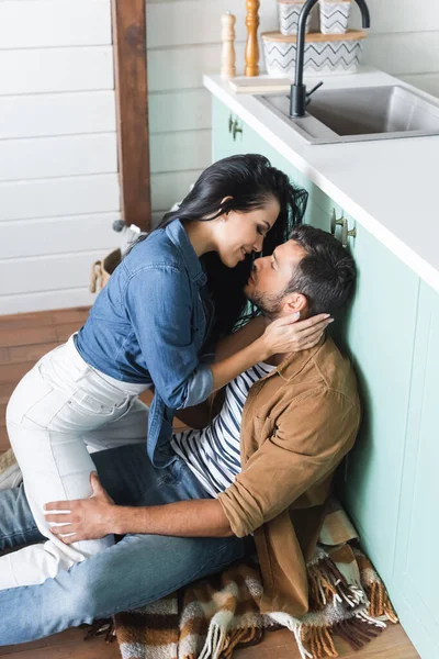 Чувственная брюнетка женщина целует парня сидя на полу на кухне — стоковое фото