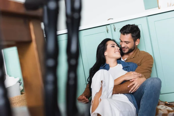 Junger lächelnder Mann umarmt glückliche Freundin, während er auf dem Boden sitzt, verschwommener Vordergrund — Stockfoto