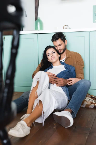 Young man embracing happy girlfriend while sitting on floor in kitchen, blurred foreground — Stock Photo