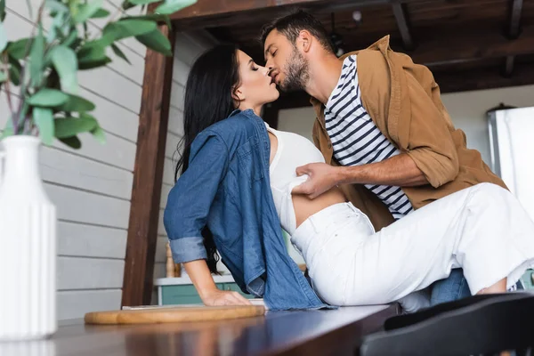 Jeune homme se déshabiller et embrasser séduisante petite amie assise sur la table dans la cuisine, flou au premier plan — Photo de stock