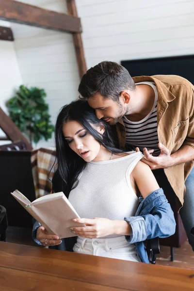 Young man seducing sexy brunette girlfriend reading book at home — Stock Photo