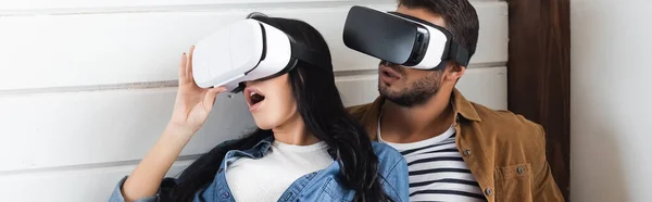 Mujer asombrada tocando auriculares vr mientras juega con el novio en casa, pancarta - foto de stock