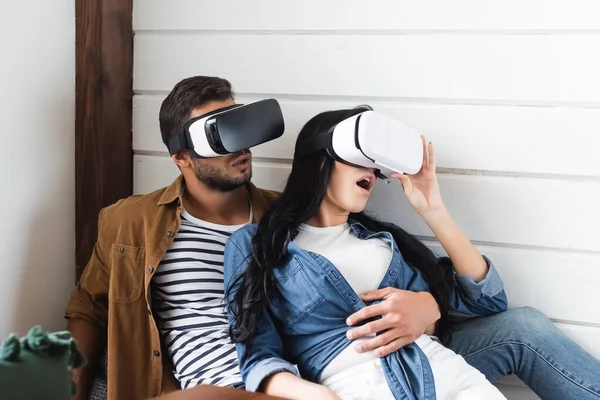 Mujer sorprendida tocando auriculares vr mientras juega junto con el novio en casa - foto de stock