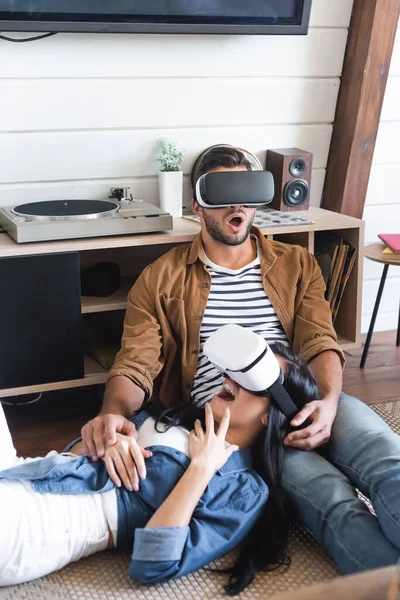 Pareja emocionada en elegantes juegos de ropa casual en auriculares vr en el suelo en casa - foto de stock