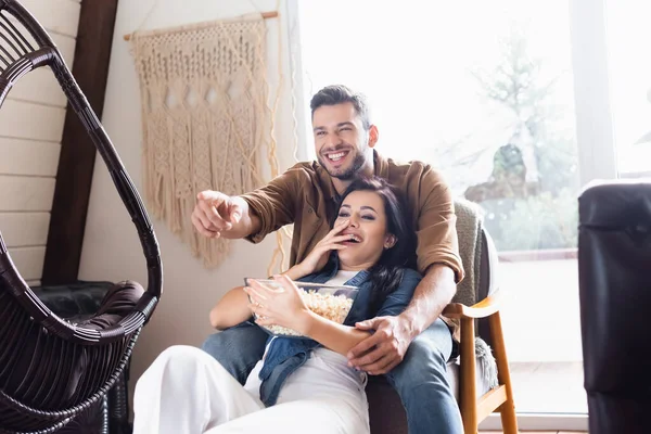 Cheerful man pointing with finger while watching tv with laughing girlfriend — Stock Photo