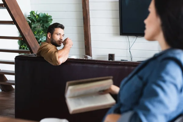 Junger Mann sieht Frau mit Buch im verschwommenen Vordergrund an — Stockfoto