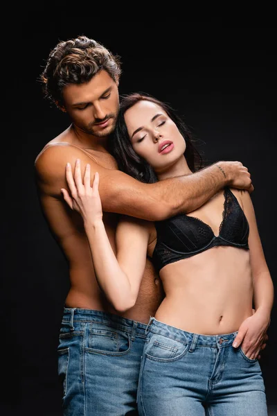 Joven hombre sin camisa abrazando a mujer seductora en sujetador y jeans aislados en negro - foto de stock